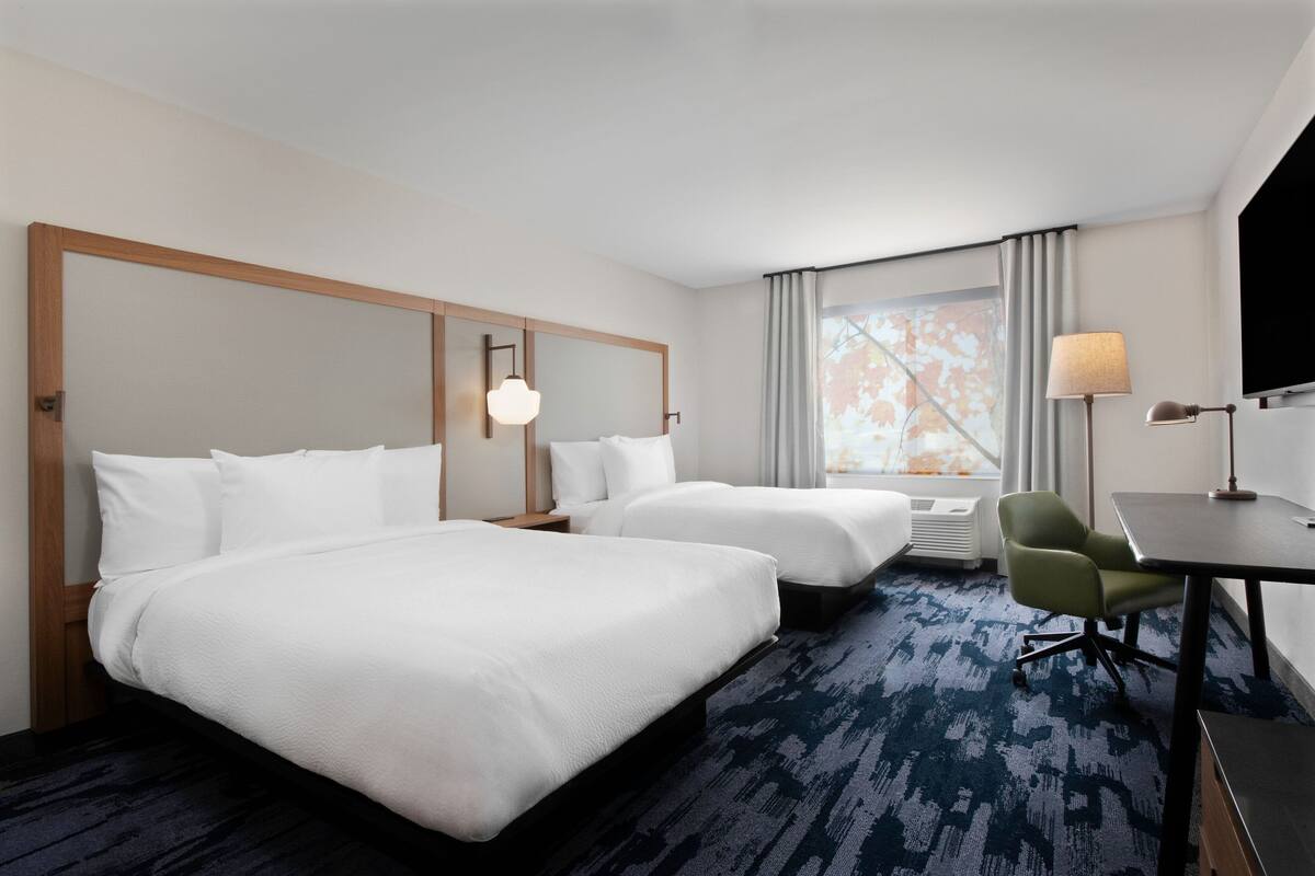 A hotel room with two beds, a desk, a chair, a TV, and a large window with curtains.