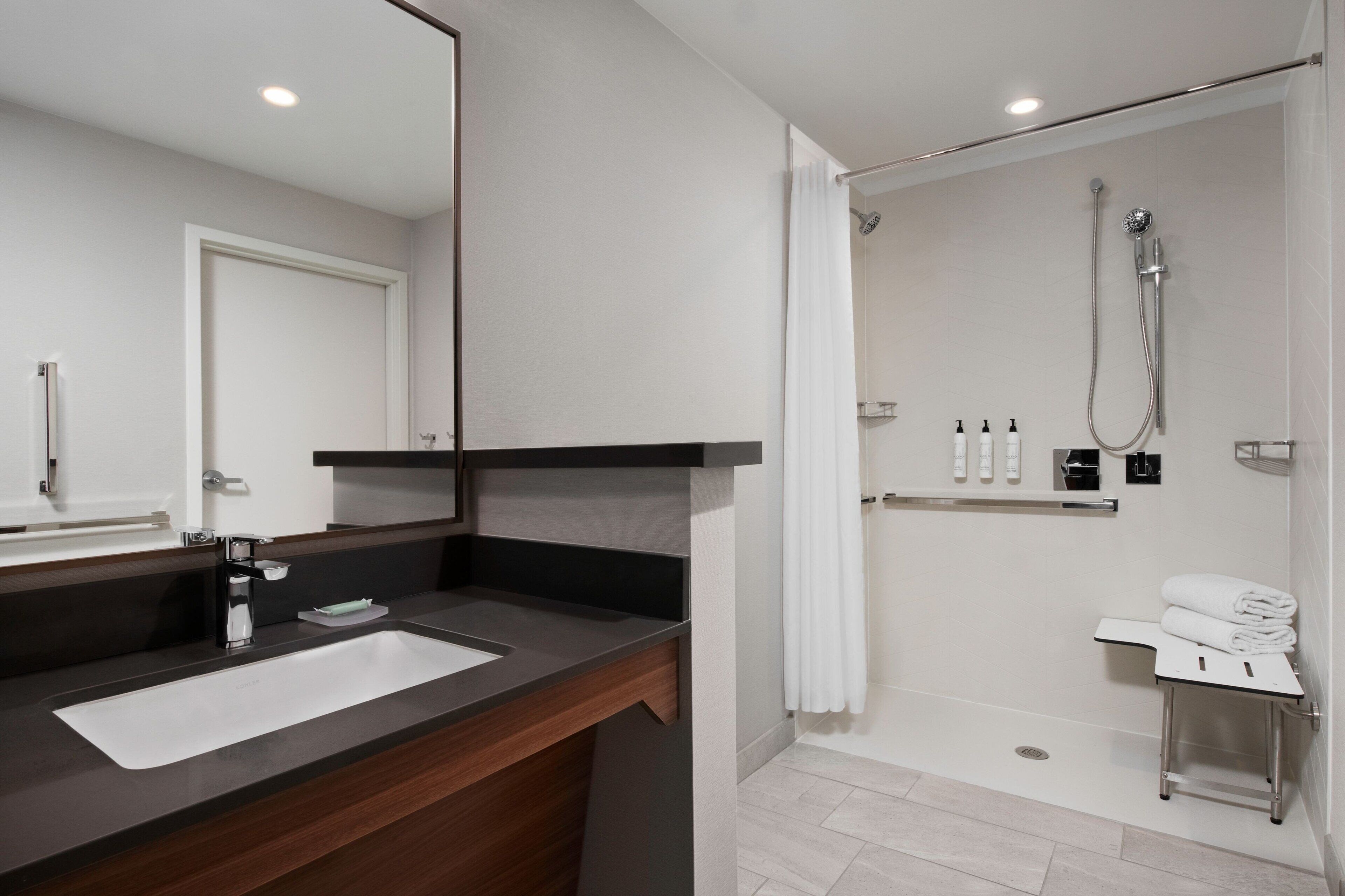A modern bathroom with a large mirror, a sink, a shower, and a small table with towels.