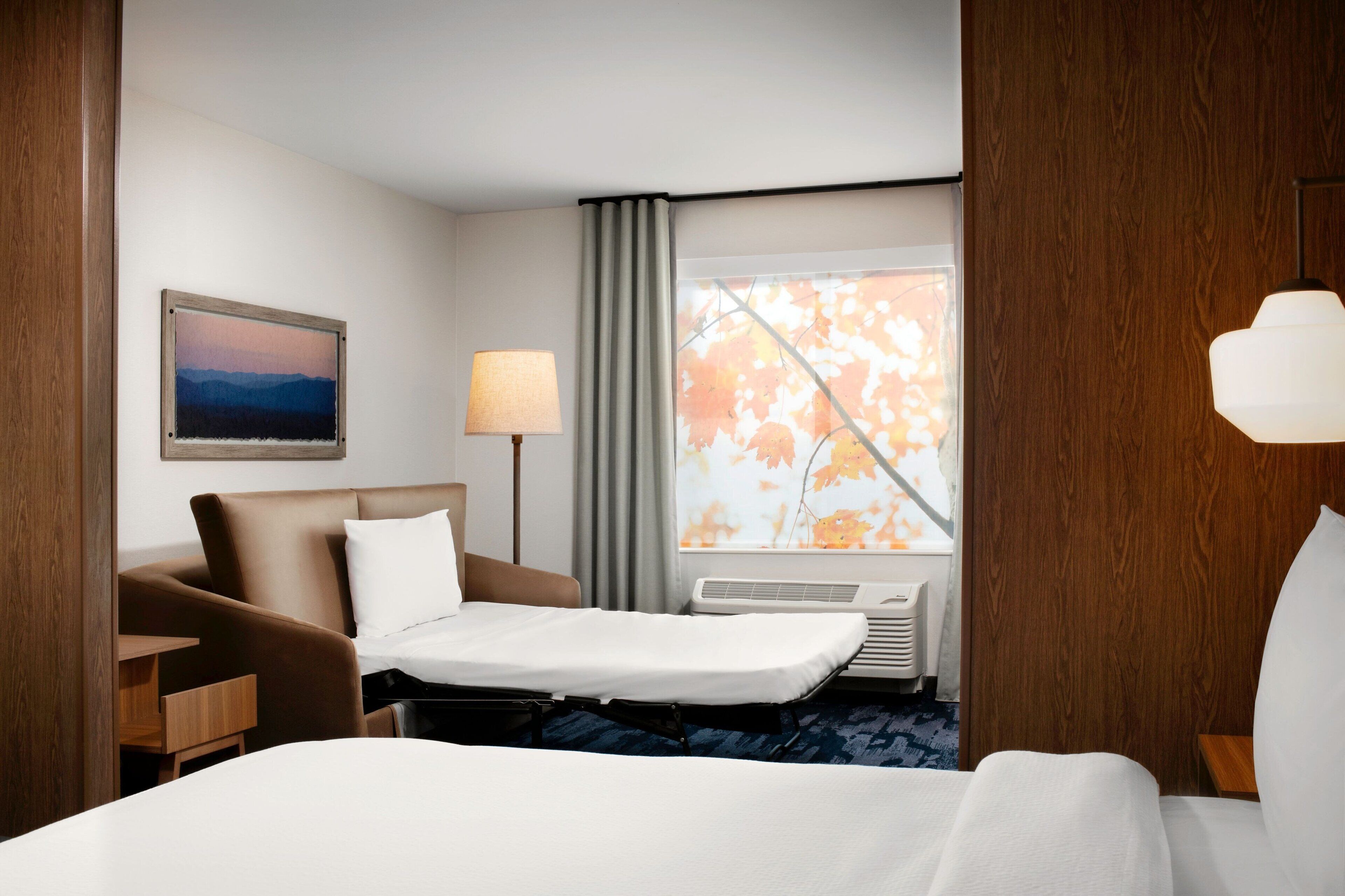 A hotel room with a bed, a desk, a chair, a lamp, a window with curtains, and a painting on the wall.