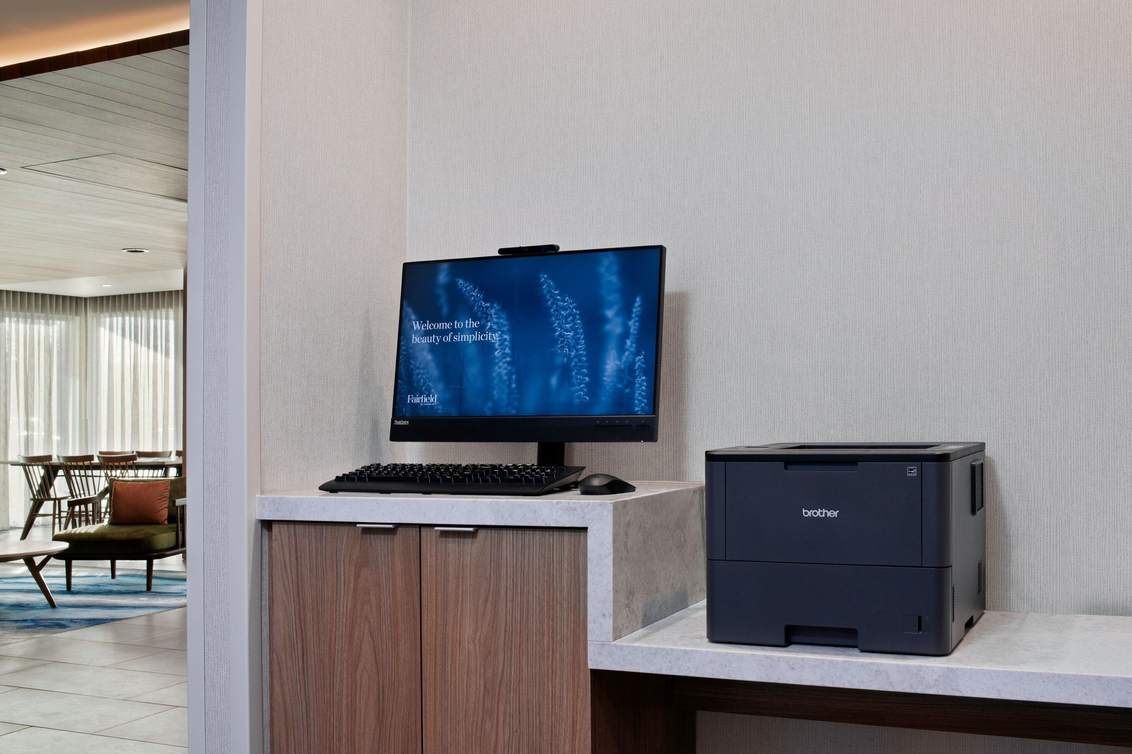 A modern hotel room with a desk featuring a Brother printer, a keyboard, and a monitor displaying a welcome message.