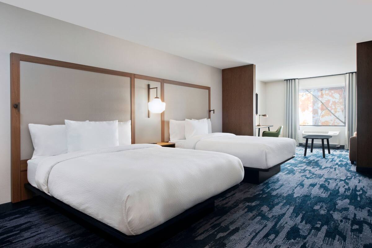 A hotel room with two beds, a small table, a chair, and a window with curtains.