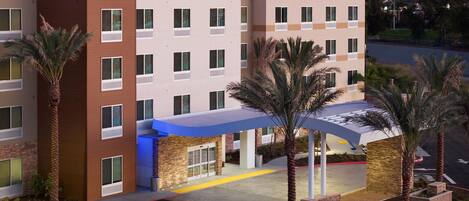 A multi-story hotel with a modern design, featuring a prominent entrance with a blue awning and palm trees.