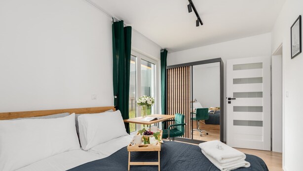 Una habitación de hotel moderna con una cama grande, una mesita de noche de madera con una bandeja de desayuno, una silla verde y vistas a la ciudad desde la ventana.