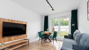 Una sala de estar moderna con un televisor de pantalla plana montado en un panel de madera, un sofá gris, una mesa de comedor con sillas verdes y un ventanal con cortinas.