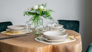 Una mesa de madera redonda con platos, tazones y vasos blancos, junto a una jarra de vidrio con follaje verde y flores blancas.