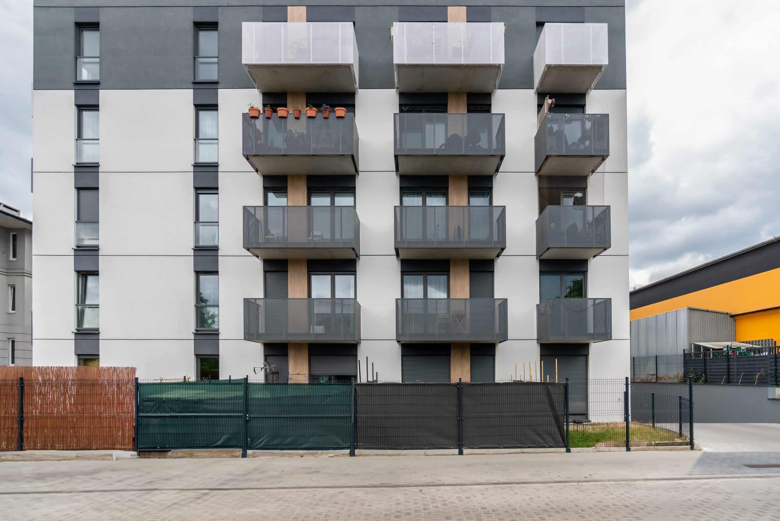 A multi-story residential building with balconies, a green fence, and a paved ground.