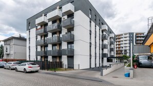 Un edificio moderno de varios pisos con balcones, un estacionamiento con automóviles y un cielo despejado.