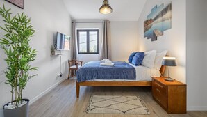 A bedroom with a bed, bedside table, lamp, TV, window, and a potted plant.