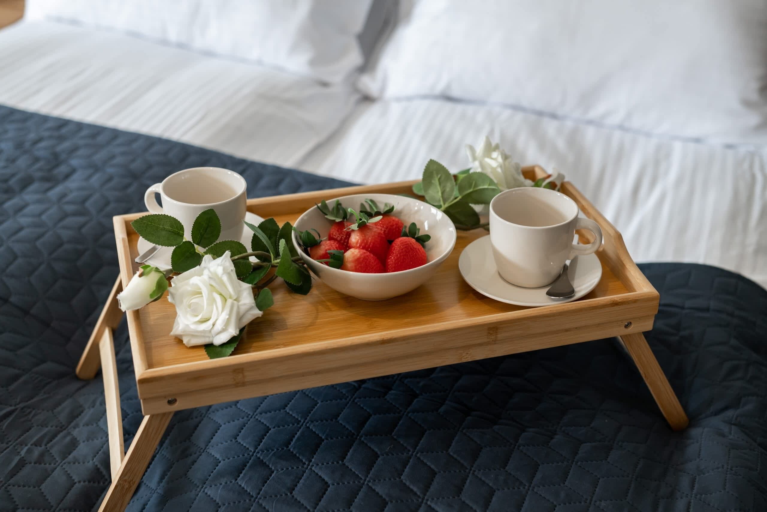 A wooden tray with two cups of coffee, a bowl of strawberries, and a white rose on a bed with a dark quilted cover.