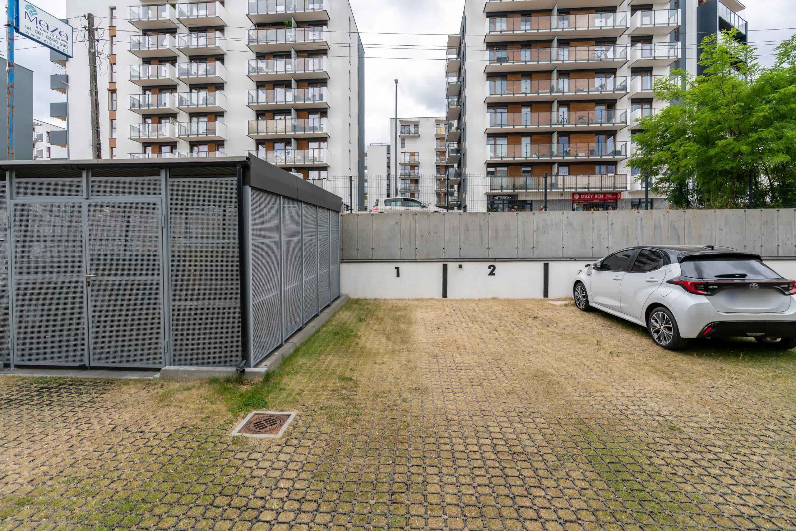 A parking area with a car, a metal fence, and a building with multiple balconies.