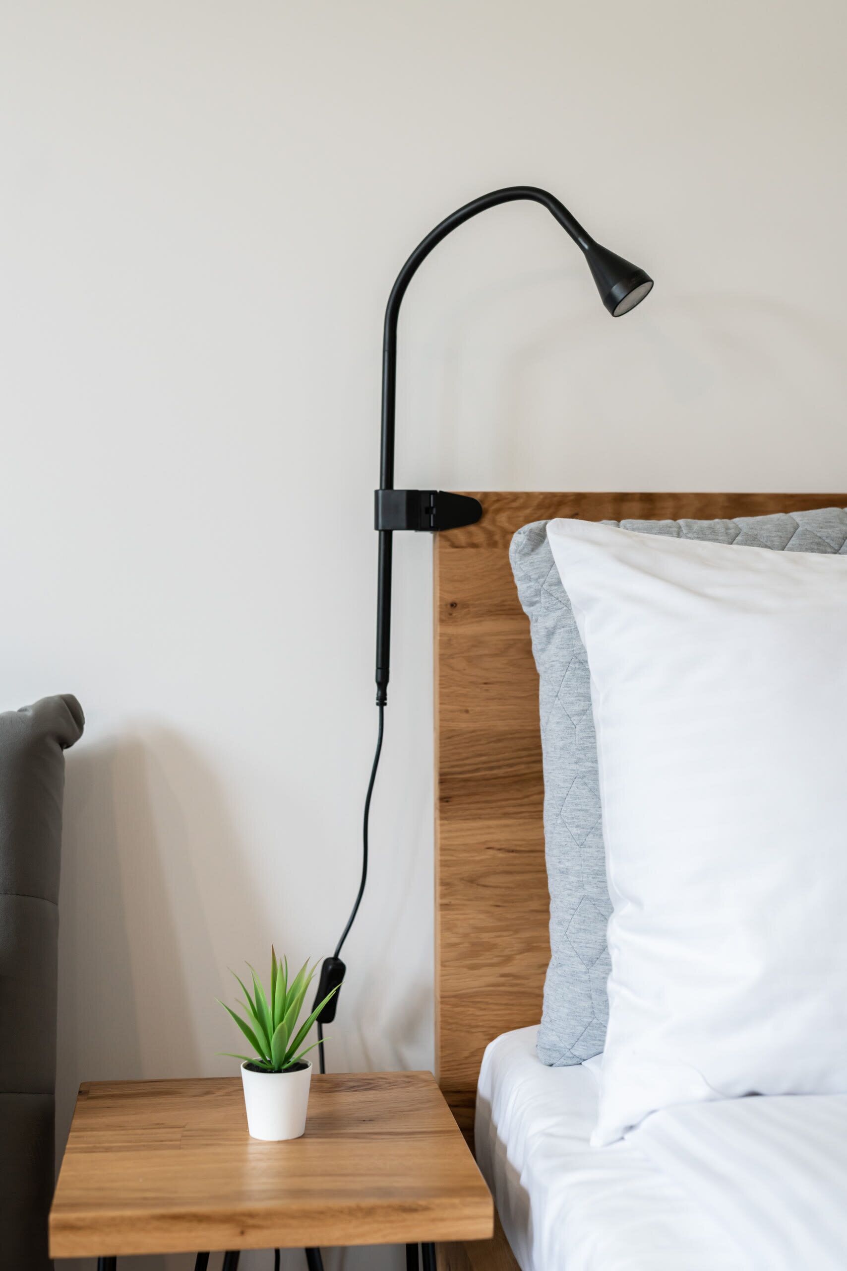 Ein Bett mit Holz-Kopfstück, ein Nachttisch mit Topfpflanze und eine schwarze Schreibtischlampe.