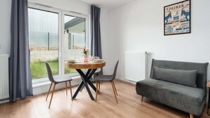 Een moderne woonkamer met een grijze bank, een ronde houten eettafel met twee stoelen en een groot raam met gordijnen.