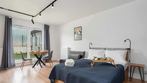 Een moderne slaapkamer met een groot bed, een klein tafeltje en een stoel. Er is een raam met gordijnen en uitzicht op de omgeving.