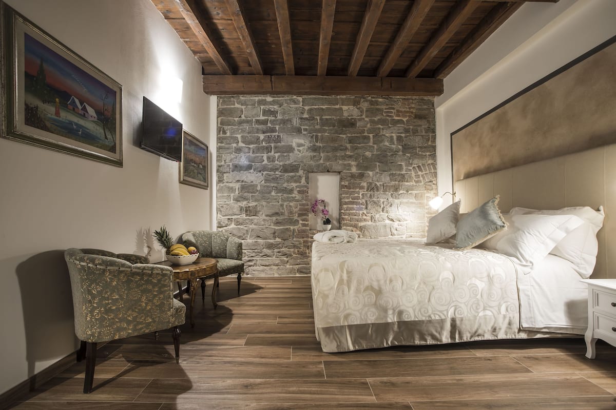 A bedroom with a stone wall, a bed with white bedding, a wooden floor, a chair, and a small table with fruit.