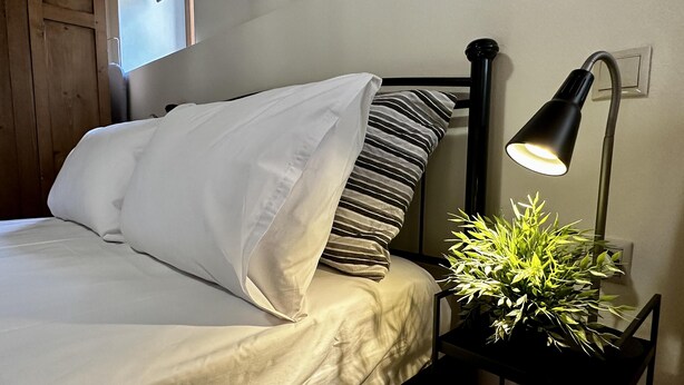 A neatly made bed with white and striped pillows, a black nightstand with a lamp, and a potted plant. - City Center 1-bed House in Heraklion (Heraklion)