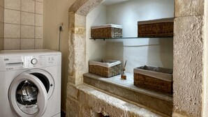 A laundry area with a front-loading washing machine, stone walls, and shelves with baskets. - City Center 1-bed House in Heraklion (Heraklion)