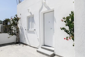 A white building with a door and a window, a small set of steps, and a wall-mounted light fixture. - Santorini Life Vibrant Studio Only 50m from the Beach (Perissa Santorini)