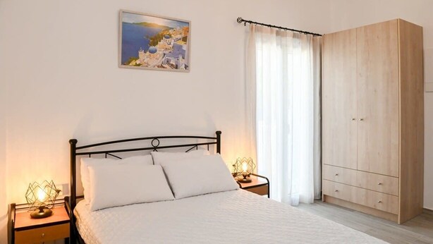 A bedroom with a bed, two bedside tables, a nightstand, a wardrobe, a painting on the wall, and a window with curtains. - Santorini Life Vibrant Studio Only 50m from the Beach (Perissa Santorini)
