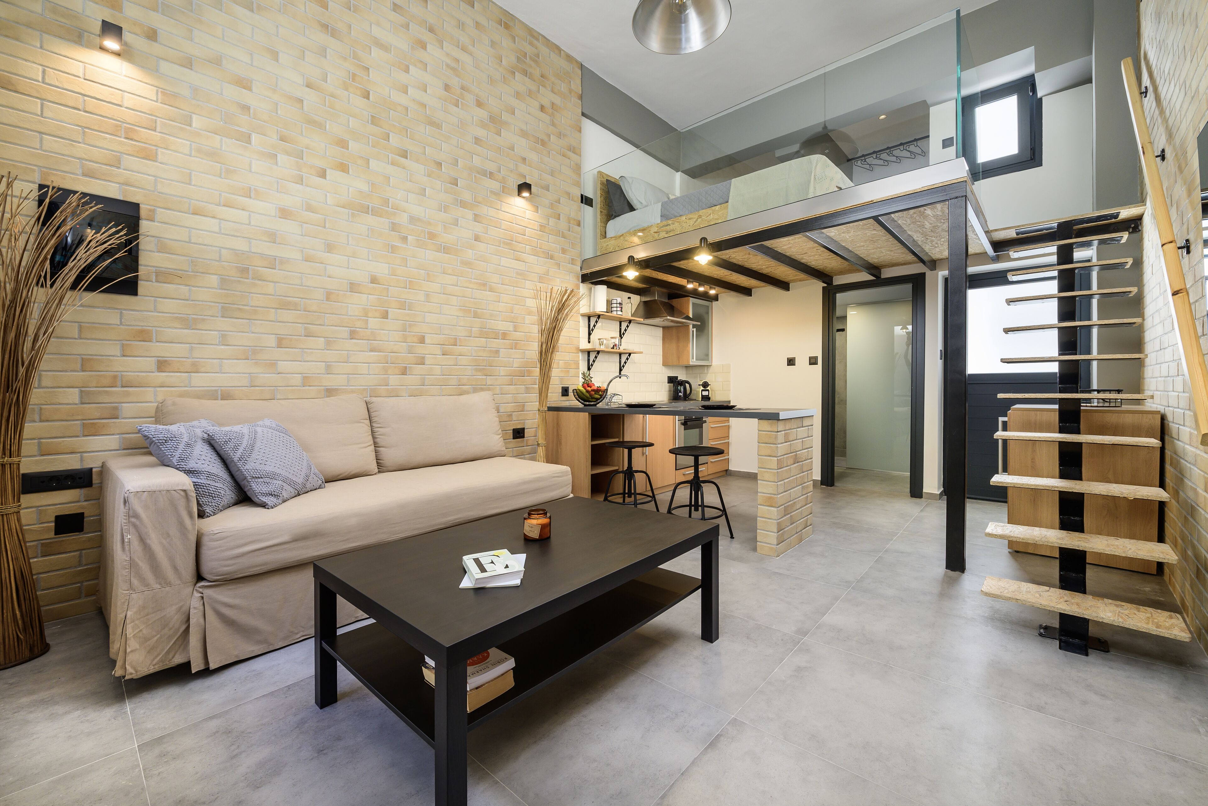 Een moderne woonkamer met een bank, een salontafel en een metselwerk muur. Er is een keukenhoek met een aanrecht en krukken.