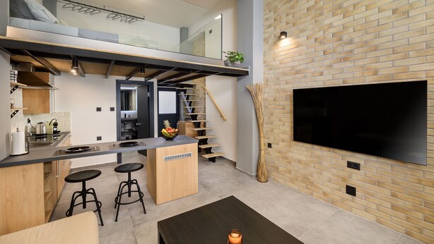 A modern kitchen with a built-in stove, a dining area with stools, a staircase, and a TV mounted on a brick wall. - Loft 2 (Kos)