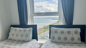 Two twin beds with matching floral-patterned bedding, a window with a view of the sea and a promenade, and blue curtains. - Ocean Wave (Bridlington)