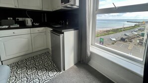 A small kitchen with white cabinets, a microwave, and a refrigerator. There is a window with a view of the sea and a parking lot. - Ocean Wave (Bridlington)