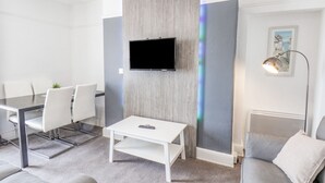 A modern living room with a flat-screen TV mounted on a wooden accent wall, a white coffee table, grey sofa, and a dining area with a glass table and white chairs. - Ocean Wave (Bridlington)