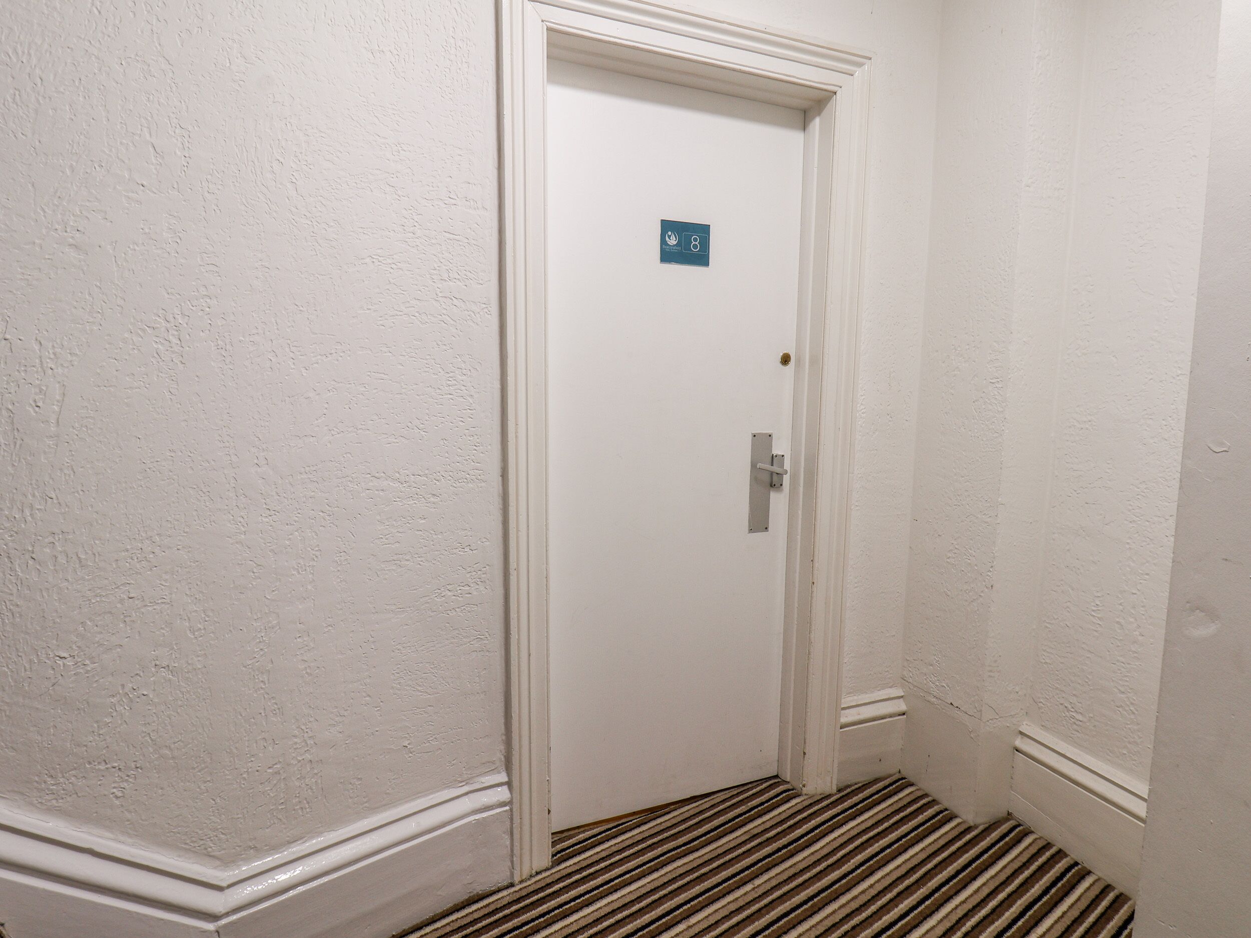 Puerta blanca con un letrero azul que indica el número de habitación 8, un picaporte metálico y un suelo cubierto de alfombra a rayas.