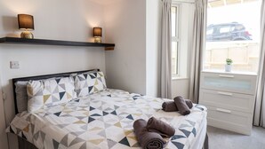 Ein Hotelzimmer mit einem Bett, einem Fenster mit Vorhängen, einem kleinen Tisch mit Pflanze und einem an der Wand befestigten Regal mit zwei Lampen.