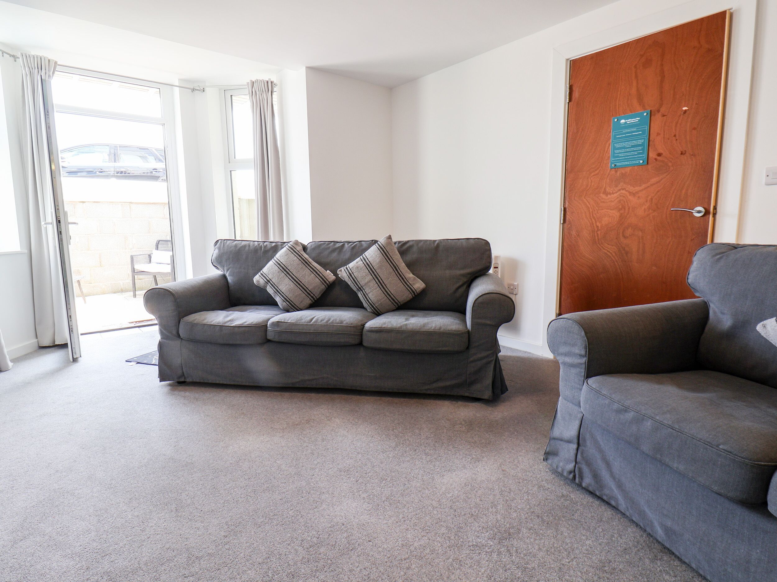 A living room with a grey sofa, two striped cushions, a wooden door, and a view through a glass door.