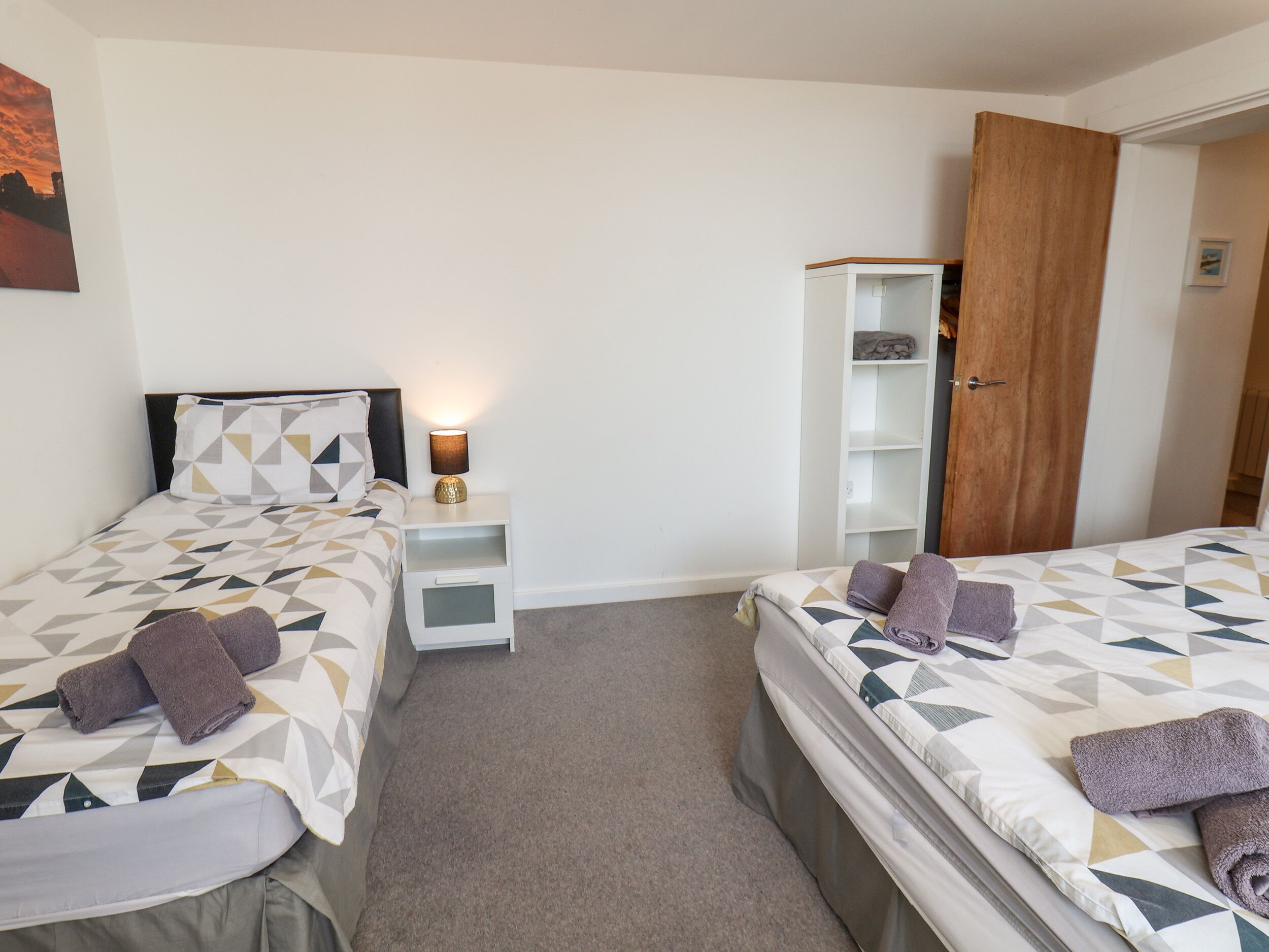 A hotel room with two beds, a nightstand with a lamp, a wardrobe, and a painting on the wall.