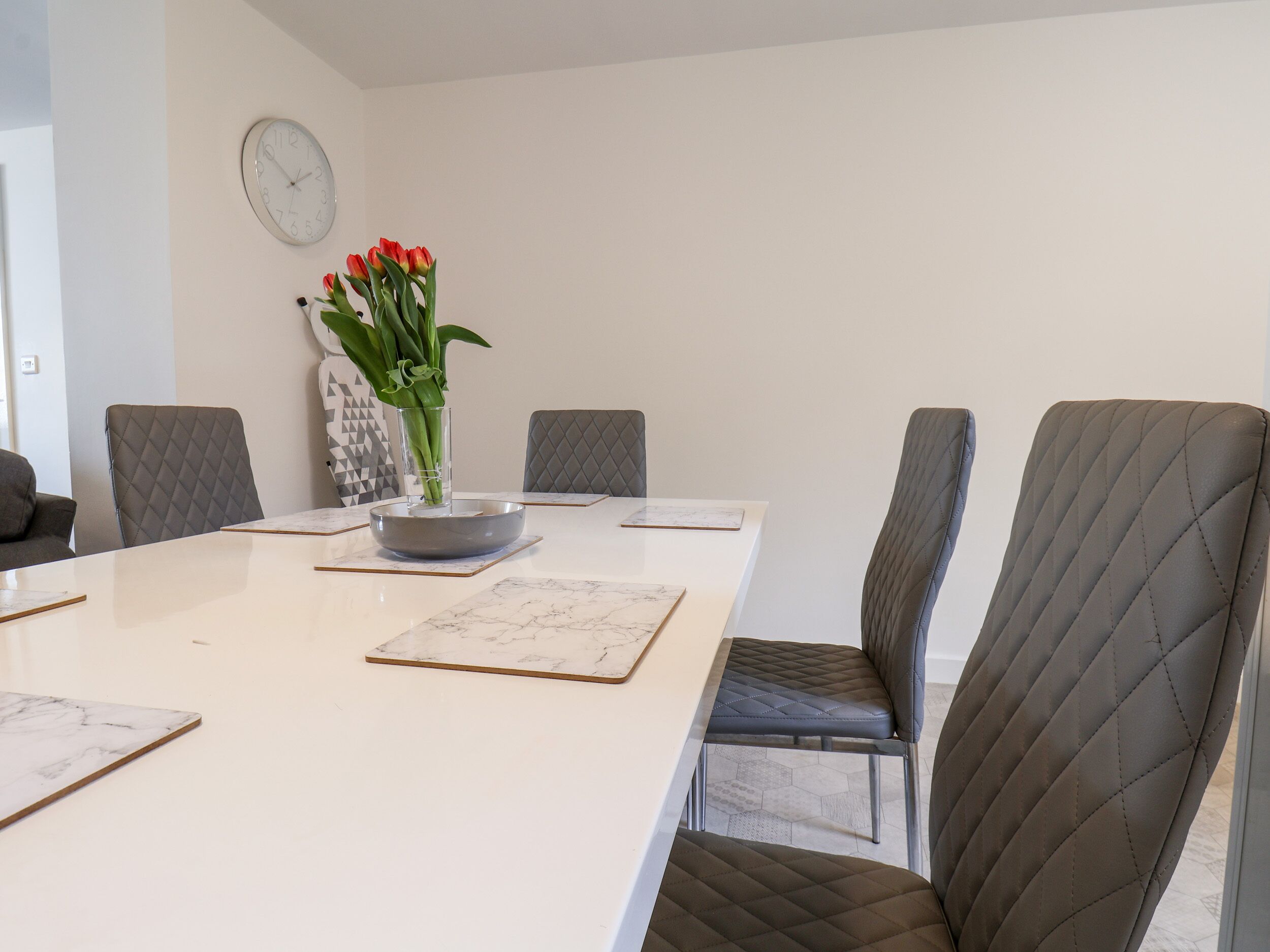 Un coin repas avec une longue table, six chaises, un vase de fleurs et une horloge au mur.