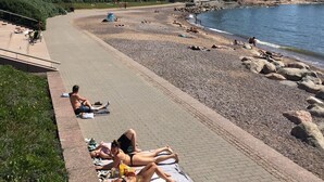 Uma passarela pavimentada ao longo de uma margem de rio, com pessoas tomando sol em toalhas.