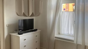 A room with a white dresser, a flat-screen TV on top, a window with white curtains, and a radiator below. - 1 Bedroom Apartment in Bonn (Bonn)