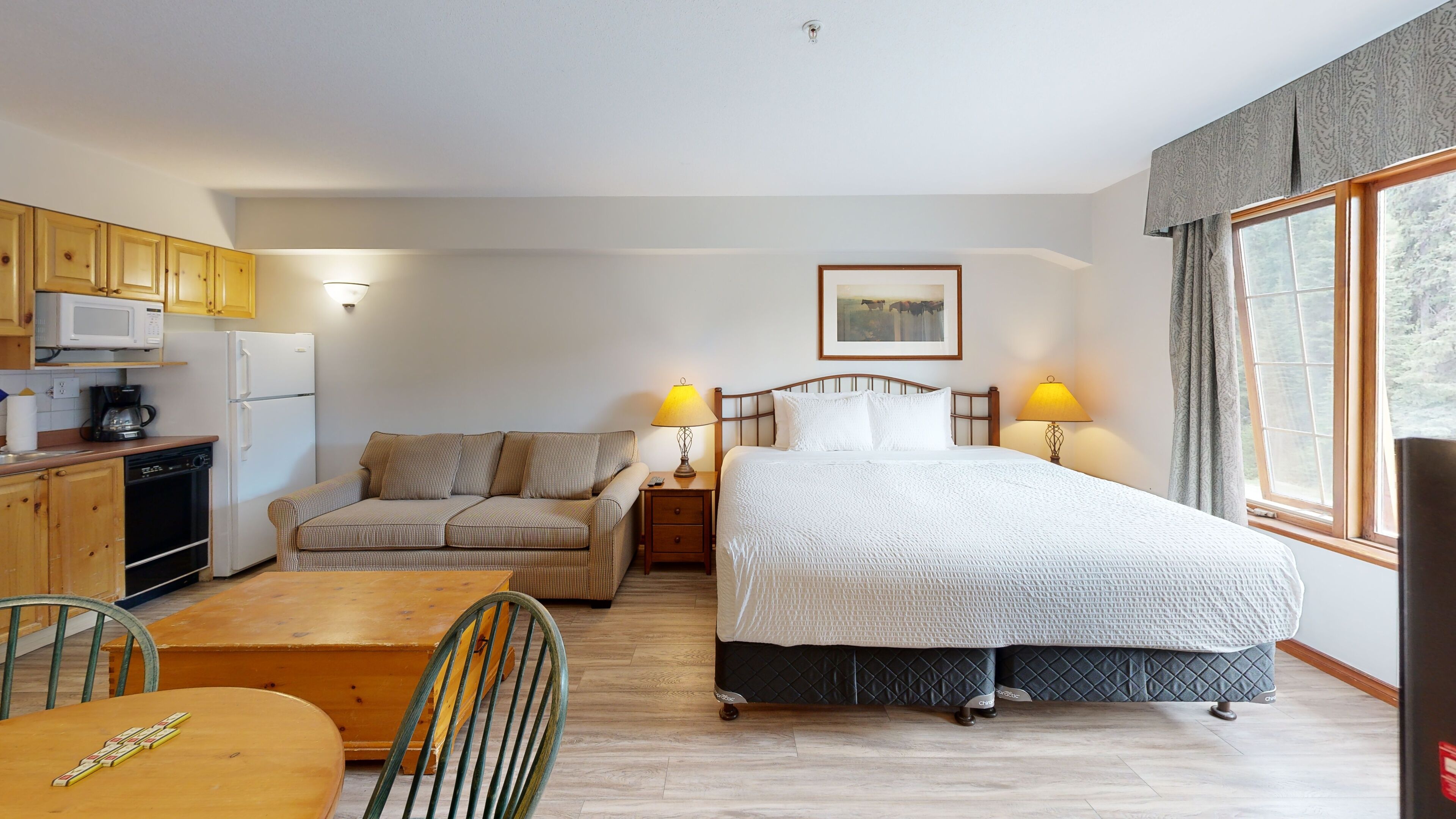 A bedroom with a bed, bedside lamps, a sofa, a dining table, and a kitchen area with a microwave and refrigerator.