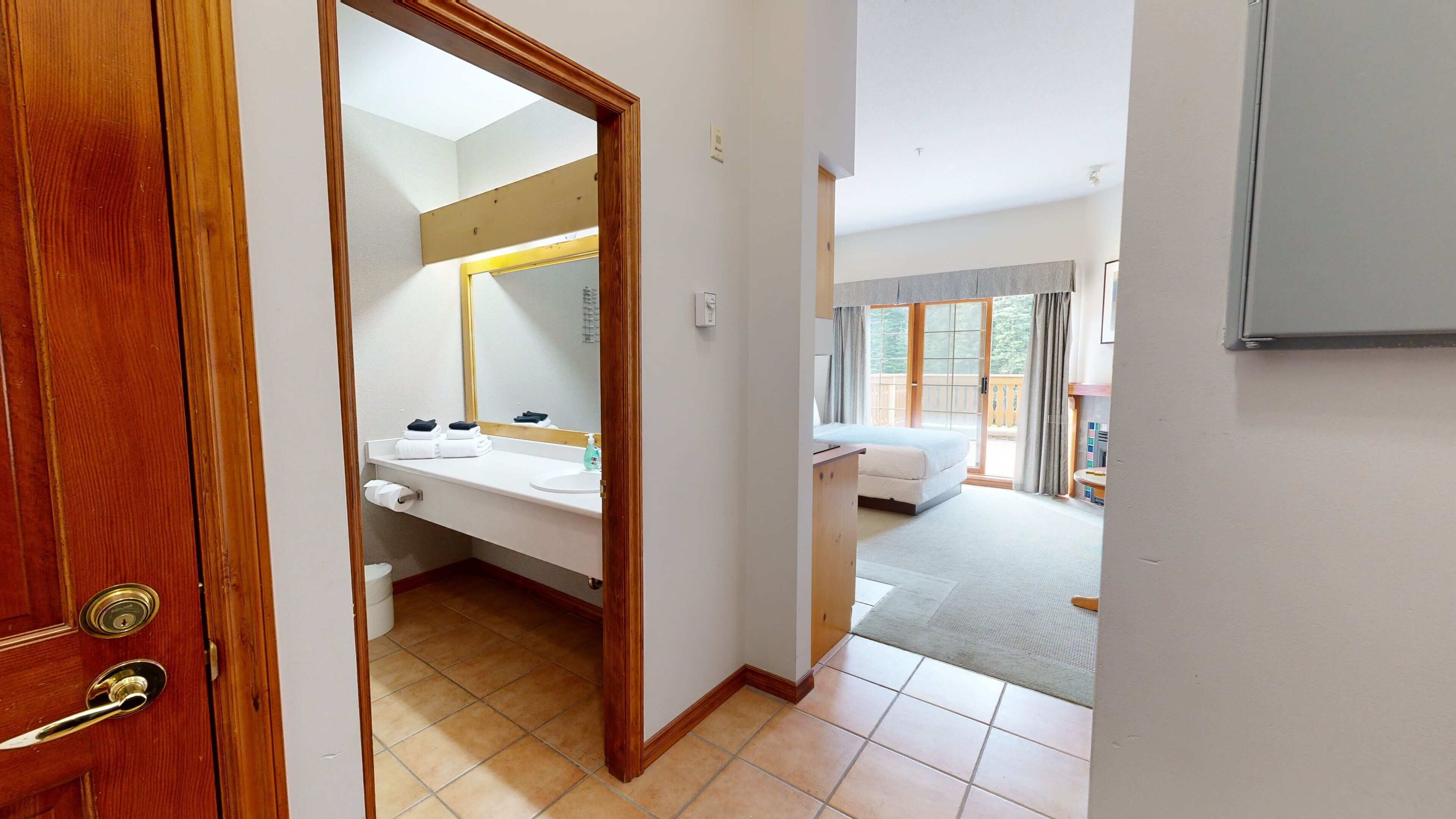 Una camera d'albergo con una porta in legno, un bagno con lavandino e specchio, un letto e vista all'esterno tramite una porta scorrevole.