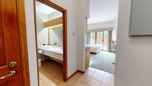 Habitación de hotel con puerta de madera, baño con lavamanos y espejo, cama y vista al exterior por una puerta corredera.
