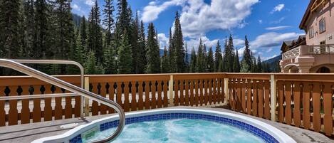 A hot tub with a wooden deck and metal railing, surrounded by a forest and a building with a balcony.