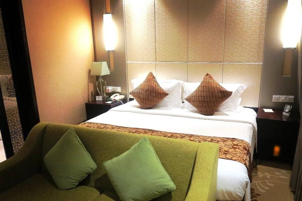 Quarto de hotel com uma cama, sofá verde e duas mesinhas de cabeceira com luminárias.