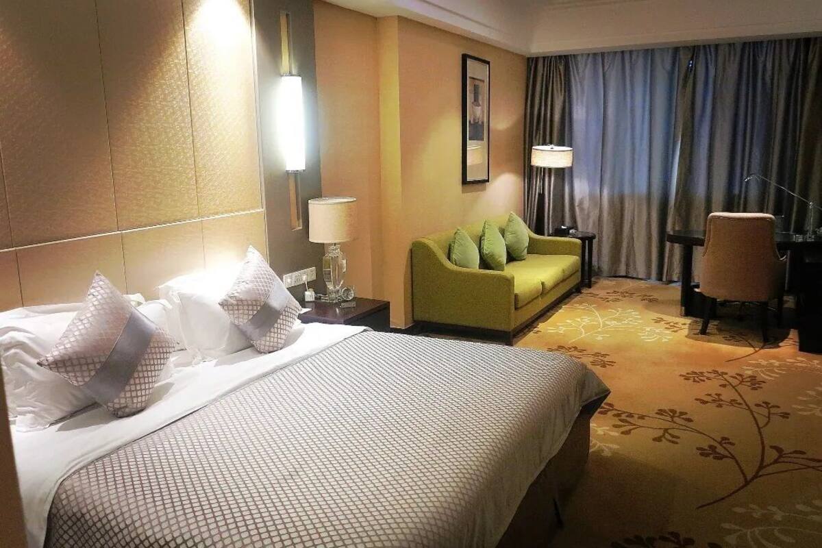 Quarto de hotel com cama, sofá verde, escrivaninha com cadeira e janela grande com cortinas.
