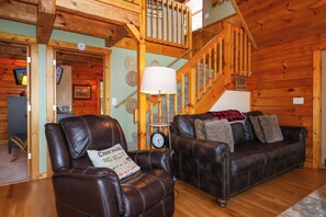 L’intérieur d’un chalet accueillant, avec un fauteuil en cuir, un escalier en bois et un canapé recouvert d’une couverture.