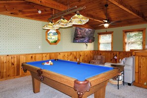 A pool table in a room with wooden walls and ceiling, a mounted television, and a ceiling fan.