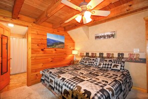 Une pièce en bois avec un lit, un ventilateur de plafond, un tableau représentant des montagnes et un mur où sont accrochées des bottes.