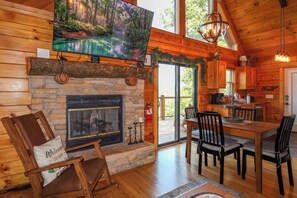 L’intérieur chaleureux d’un chalet avec une cheminée en pierre, une table à manger en bois et un téléviseur fixé au-dessus de la cheminée.