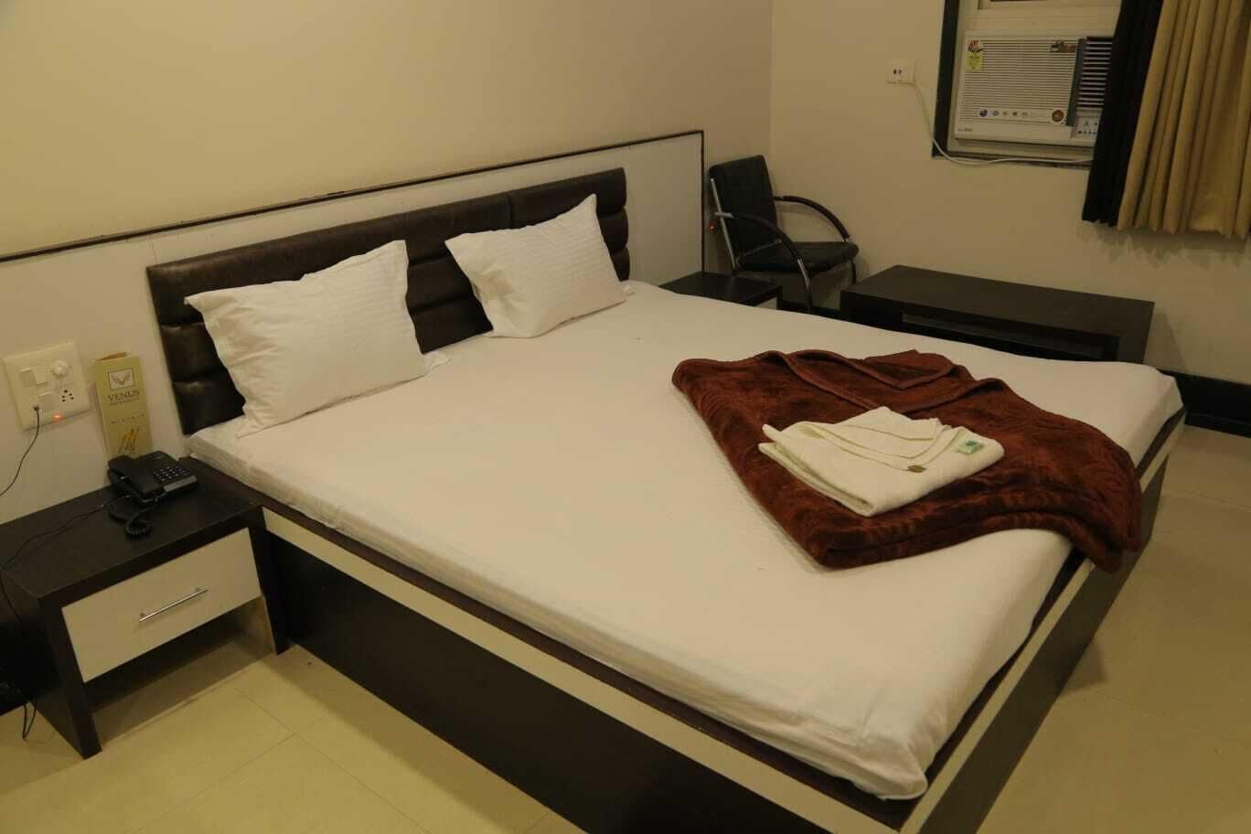 A hotel room with a bed, a bedside table with a phone, a chair, and a wall-mounted air conditioner.