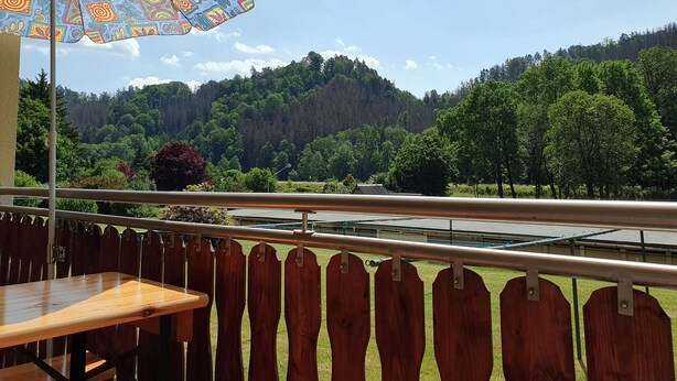 Un balcon avec une table en bois et un parasol coloré, donnant sur un paysage verdoyant et luxuriant ainsi que sur une rivière.