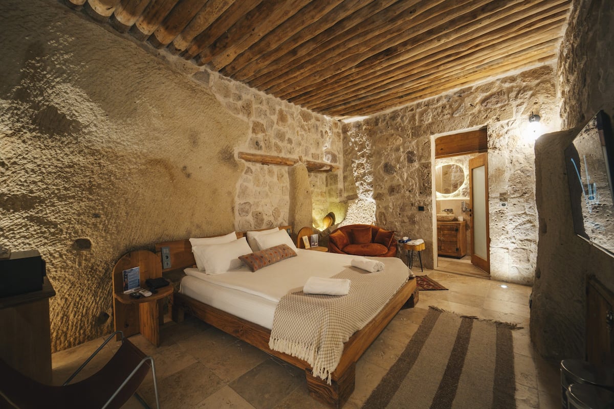 Un dormitorio rústico con paredes de piedra, una cama de madera, una mesita de noche, una silla y una pequeña mesa con lámpara.