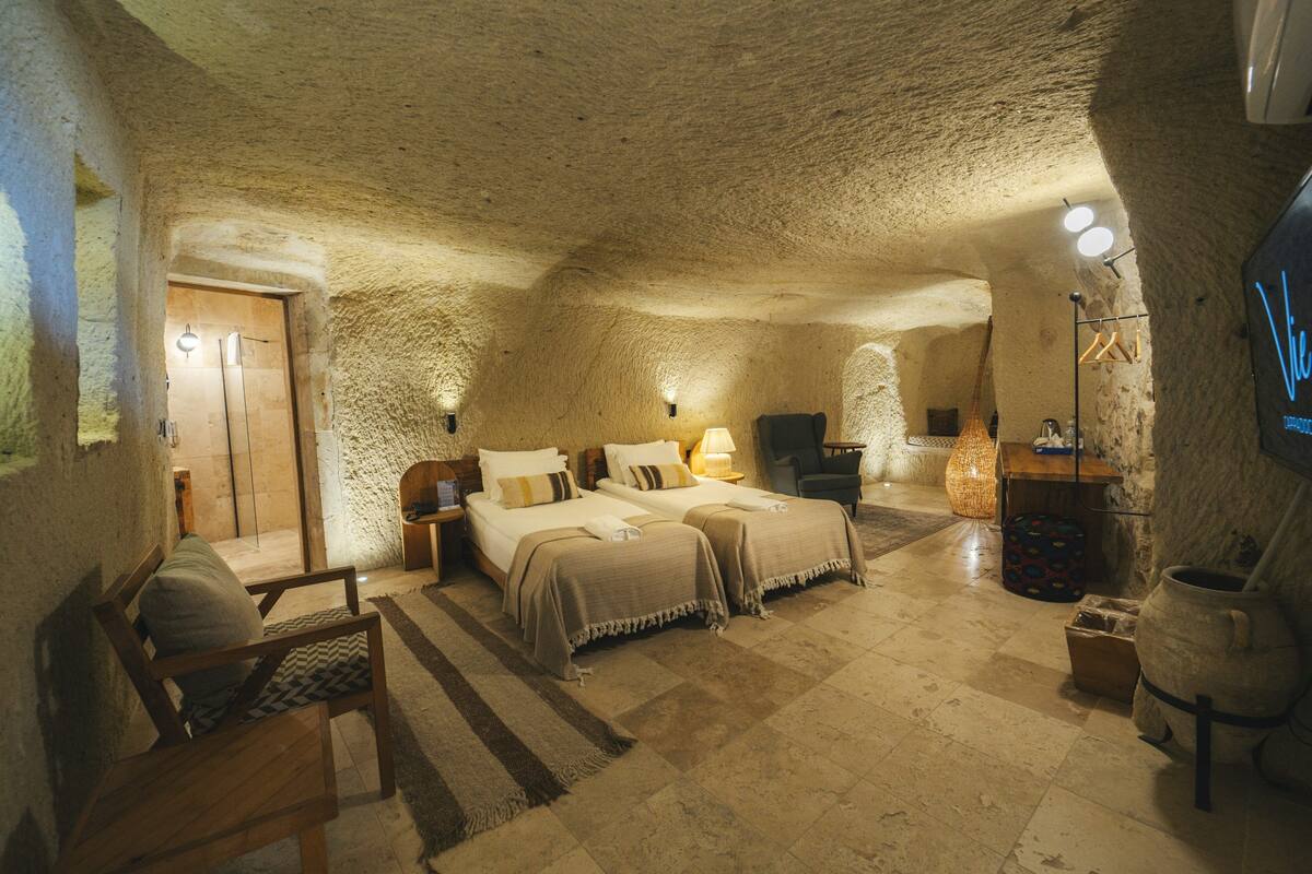 Una habitación acogedora de estilo cueva con dos camas, una silla de madera, una mesita pequeña y una gran maceta de piedra.