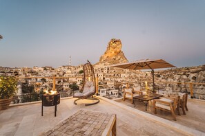 Terraza en la azotea con una silla colgante, una mesa y una sombrilla, con vistas a un paisaje urbano histórico y una imponente formación rocosa.