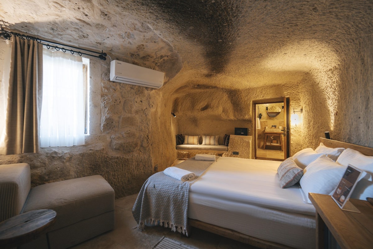Un dormitorio acogedor con paredes de piedra, una cama grande, una mesita de noche y un pequeño rincón de descanso.